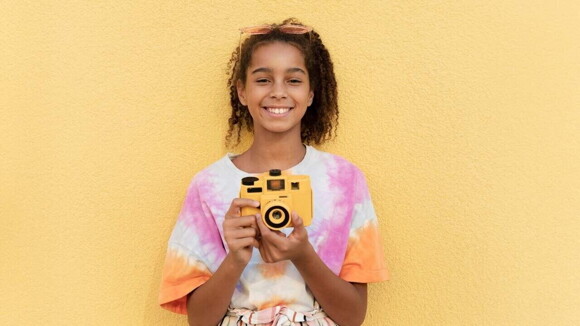 Menina segurando câmera fotográfica e sorrindo em frente a uma parede amarela.