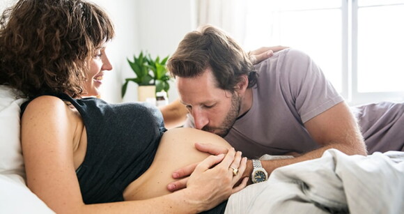 Mulher grávida sorrindo, deitada na cama, enquanto homem beija a barriga dela