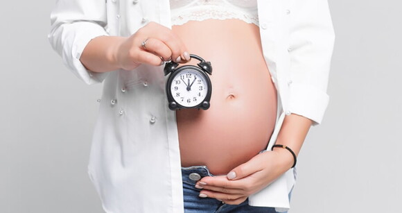 Mulher grávida com a mão sob a barriga e segurando um relógio.