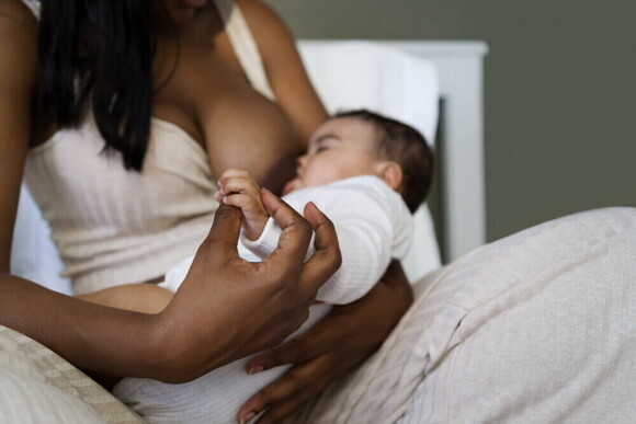 Mãe sentada, amamentando um bebê em seu colo e segurando a mão dele