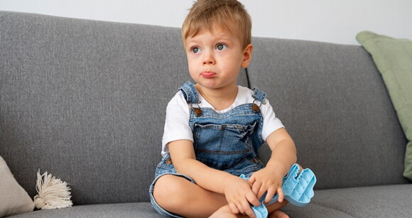 Menino usando macacão jeans sentado no sofá segurando peças de um brinquedo de montar.