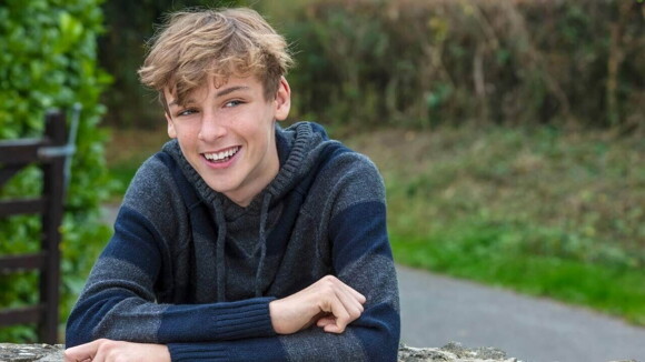 Um jovem sorrindo, sentado em uma mesa ao ar livre, vestindo uma roupa de frio.