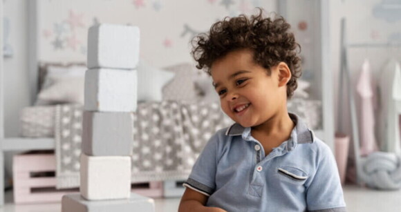 Criança sorrindo enquanto brinca com blocos empilhados, usando uma camisa azul em um ambiente infantil decorado.