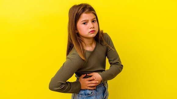 Uma menina em um fundo amarelo, com expressão de dor, segurando a região abdominal, sugerindo desconforto.