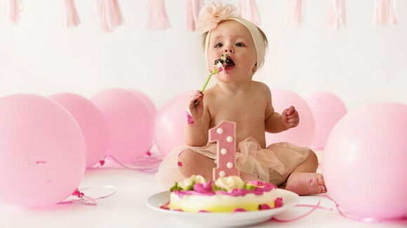 Um bebê comendo um bolo, ao lado de balões de aniversário rosa.