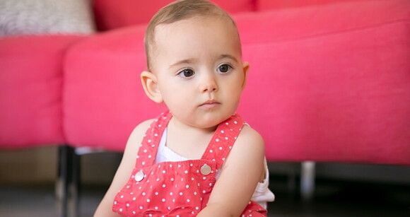 Menina de aproximadamente 1 ano sentada no chão da sala usando um vestido cor de rosa com bolinhas brancas.